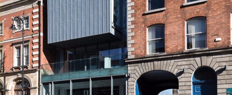 Photo of the exterior of the gallery, viewed from across Thomas Street in the Liberties, Dublin on a sunny day; the gallery is modernist in style, inserted between more traditional redbrick buildings; to the right of the gallery we see the archway of the National College of Art and Design; people are walking by