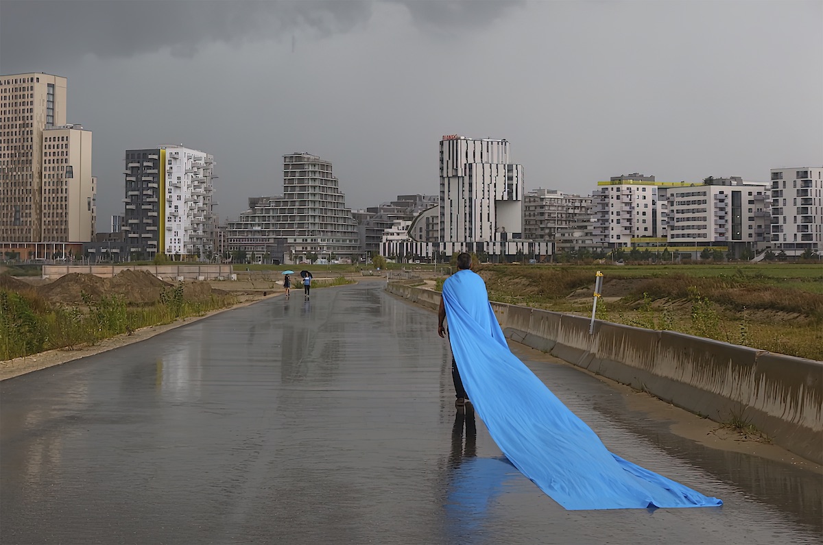Sérgio Leitão: UILLINN_THE FALLOUT | Saturday 1 April – Saturday 6 May 2023 | Uillinn: West Cork Arts Centre | Image: photo of man standing on wet road in rain, facing a series of larger modern housing developments in the distance; he's wearing a very long light-blue cape or train 