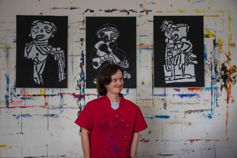 Photo of Ailbhe Barrett © Clare Keogh | The Power of Us | Friday 31 March 2023 – Sunday 3 March 2024 | Crawford Art Gallery | Image of Ailbhe Barrett © Clare Keogh – photo of the artist in a studio setting, wearing a red sleeveless top, with artworks on the wall behind 