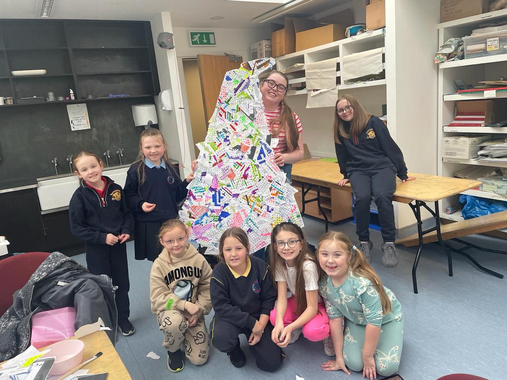 Image courtesy of the artist Emma Brennan | Dublin Learning City Festival 2023 | Wednesday 5 April – Thursday 6 April 2023 | NCAD Gallery | Image courtesy of the artist Emma Brennan – we see what is probably a national-school Art room; seven pupils are present, plus an older person holding up a probable artwork in the shape of a flat Christmas tree and covered in stuck-on coloured bits of paper with writing (?) and marks on them 