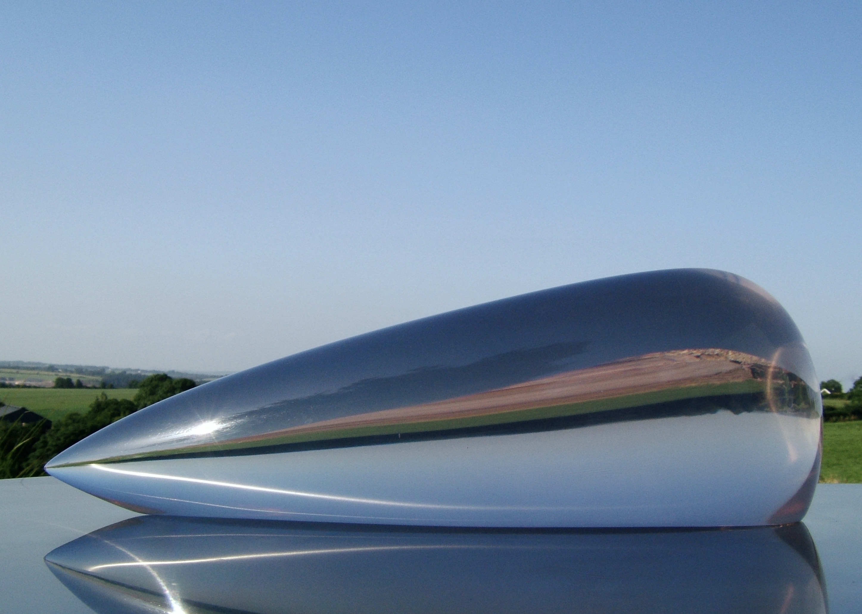 Eilis O’Connell: A Family of Things | Saturday 17 June – Saturday 30 September 2023 | F.E. McWilliam Gallery | Image: photo of a highly polished lozenge-shaped metal object, probably of significant size, on a reflective plinth; the surrounding countryside is mirrored back, distorted · \