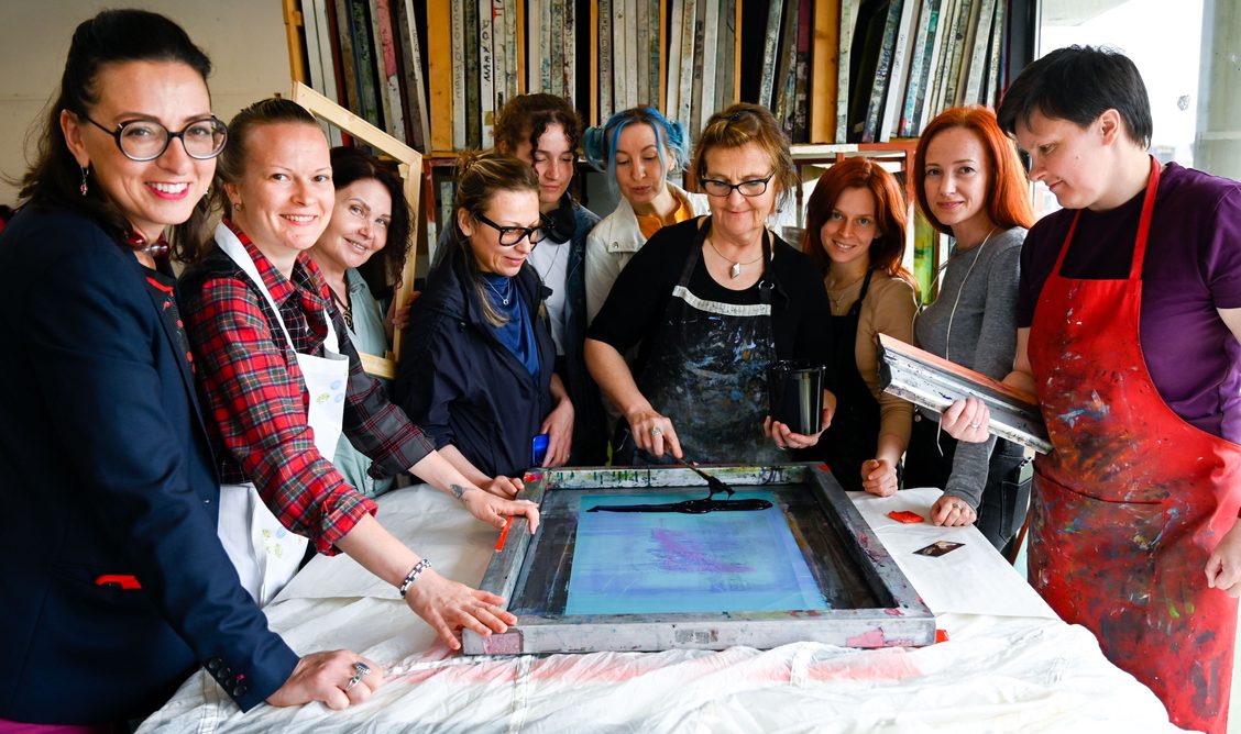 InFlight_OnLanding | Tuesday 20 June – Tuesday 27 June 2023 | | Image: photo of artist Varvara Keidan Shavrova and ten refugees gathered around as ink is applied to a silkscreen mesh for printing onto what appear to be white fabric 