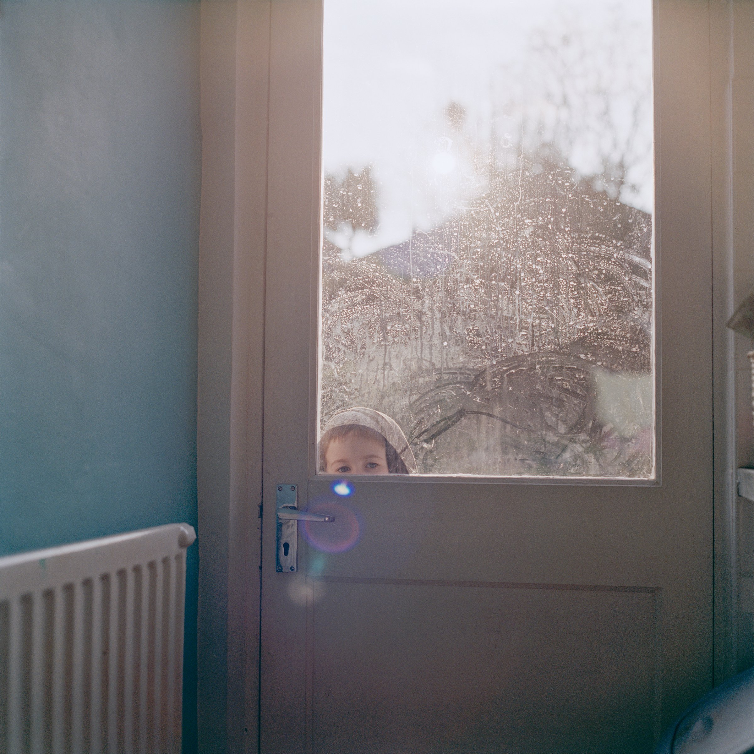 from the series Domestic Drift; © Clare Gallagher | No Place Like Home: The Domestic in Irish Photography | Thursday 20 July – Saturday 2 September 2023 | Photo Museum Ireland | Image: from the series Domestic Drift, © Clare Gallagher – from inside, we see what is probably the back door of a house; there may be rain on the window of the door, though there’s a hazy sun visible; we just see the eyes and top of the head of a child looking in through that window 