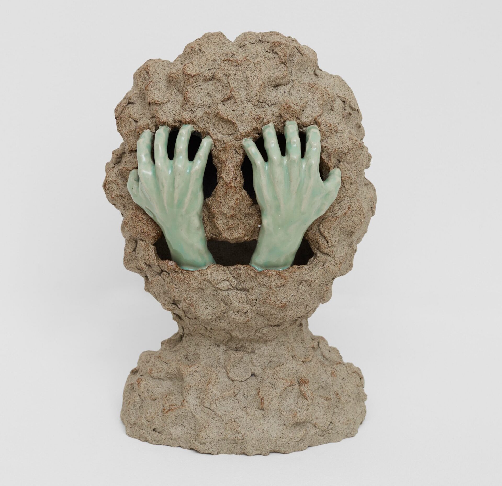 William Cobbing: Grapnel, 2023 | CLAY Festival | Friday 25 August – Sunday 27 August 2023 | National Sculpture Factory | Image: William Cobbing: Grapnel, 2023 – photo of a clay sculpture; we see a very lumpy round clay head, a bit reminiscent of the Cookie Monster, with large deep holes for the round eyes and a wide horizontal mouth; from the mouth emerges a pair of realistic hands glazed pale green on white; the hands, presumably from the same hidden body, look as though they are clawing at the eyes / eye sockets 