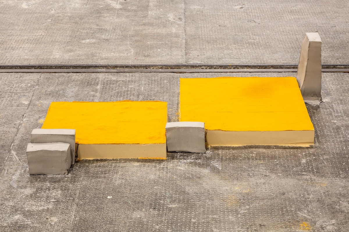 Iza Tarasewicz, Yellow Coal, 2016-2022. Courtesy of the artist and Gunia Nowik Gallery. | EVA at LCGA | Thursday 31 August – Sunday 29 October 2023 | Limerick City Gallery | Image: Iza Tarasewicz, Yellow Coal, 2016-2022. Courtesy of the artist and Gunia Nowik Gallery. | photo of what looks like an installation in a public place; there’s a textured concrete surface crossed horizontally across the photo by what looks like a single tram track; between us and the track we see two slabs of unknown material; each slab is a strong yellow on top, a creamier yellow on the sides, a bit like a cheesecake; the two slabs are not particularly aligned with each other; their dimensions are possibly around 50 to 70cm squared, and about 10cm tall; there are four other smaller slabs, grey, on their sides; one’s thicker towards the bottom. 