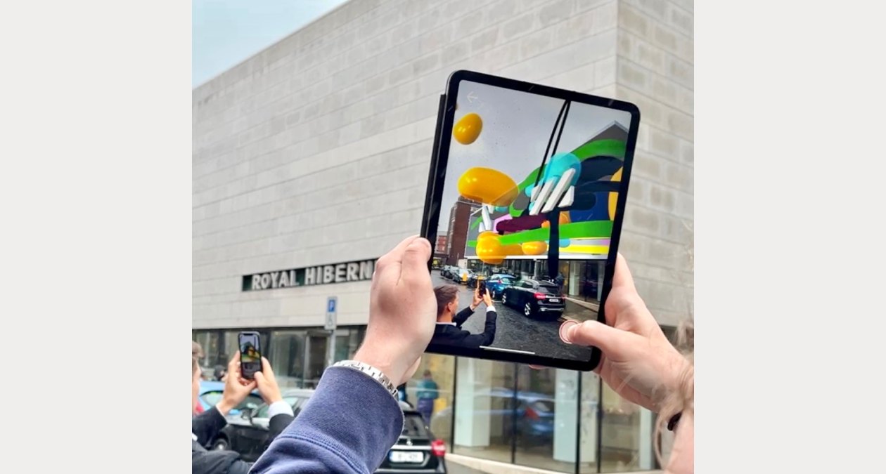 Maser: Around the Block | Friday 22 September – Sunday 1 October 2023 | Royal Hibernian Academy | Image: photo showing a pair of hands holding up a digital tablet on which we see an augmented-reality version of the façade of the RHA, with the surrounding street and people unaltered; the AR intervention has added coloured stripes and other 2D and 3D motifs, and floating balloon-like shapes in vivid orange 