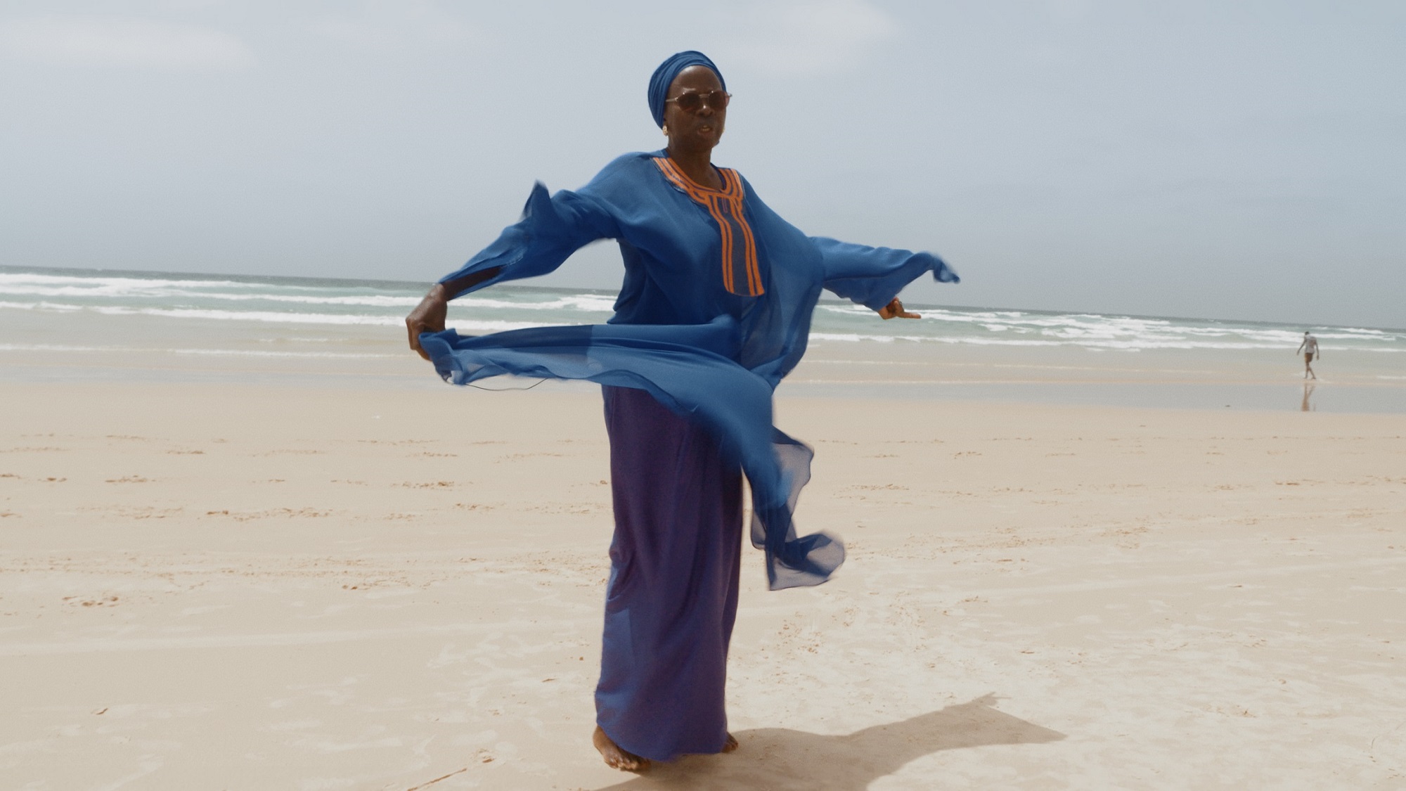 still from Leviathan Cycle Episode 7: Africana, Ken Bugul & Nemo by Shezad Dawood | EARTH RISING | Thursday 21 September – Sunday 24 September 2023 | IMMA | Image: still from Leviathan Cycle Episode 7: Africana, Ken Bugul & Nemo by Shezad Dawood | someone on a beach in flowing blue, purple and orange clothing, arms outstretched; sunglasses, and face turned towards the camera; beach and sea in the background, stretching to the tilted horizon; figure walking towards the sea towards the right edge of the photo 