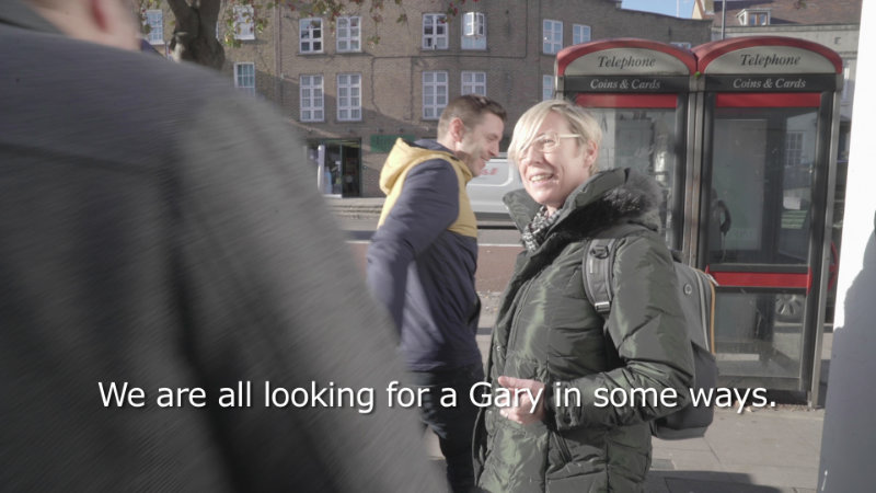 Michelle Deignan, Looking for Gary (2018) 07:20 minutes; selected by Crawford Art Gallery, Cork | Artists’ Film International screening programme | Friday 10 November – Sunday 19 November 2023 | Crawford Art Gallery | Image: Michelle Deignan, Looking for Gary (2018) 07:20 minutes; selected by Crawford Art Gallery, Cork | still from film / video taken somewhere, presumably, in the UK; there are city buildings in the background, not too high, then nearer to us two telephone kiosks with red, white and black livery, then someone passing from left to right, then closer still another person, blond-haired and wearing a rucksack (probably), who is turning to someone of whom we can only see a bit of the head but mostly the right should, close to the camera at the left side of the image; a subtitle reads “We are all looking for a Gary in some ways.” 