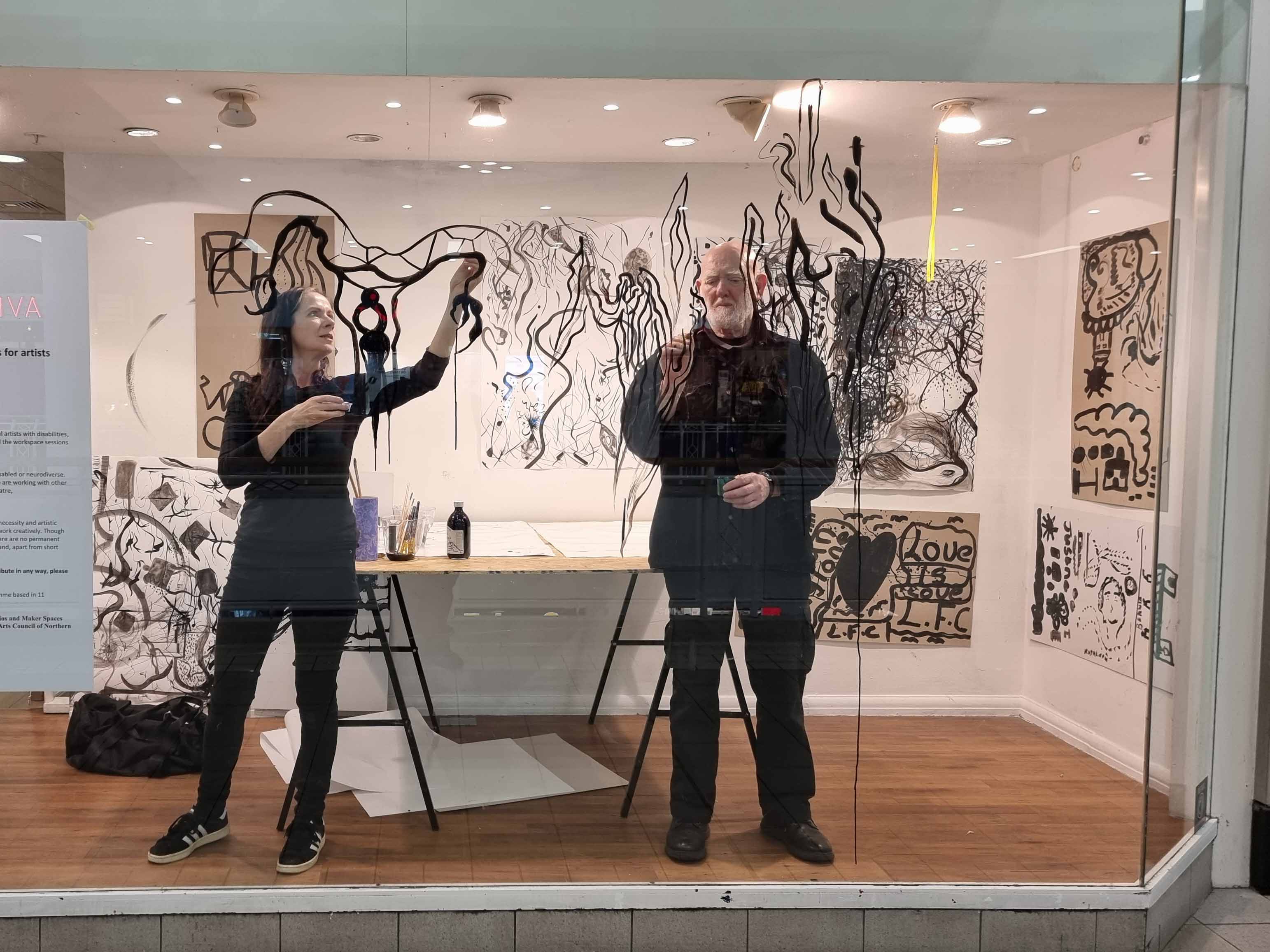 One Day Live Drawing Performance by Alastair MacLennan and Marie Phelan | Friday 12 January 2024 | The LAB | Image: photo through plate glass of the two artists drawing on the glass, with many drawings on the walls behind them and to their |eft 