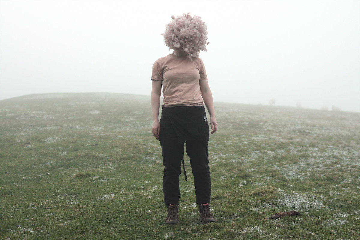 Joanna Hopkins: Fruity Bodies | Saturday 9 March – Friday 3 May 2024 | Municipal Gallery | Image: photo of female standing in a misty field; wearing black jeans and a pink T shirt, with a fluffball where the head should be 