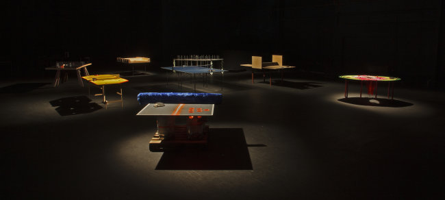 Image © Davey Moor | WIFF WAFF* is PLAYING at my GAFF | Friday 14 June – Sunday 7 July 2024 | Crawford Art Gallery | Image © Davey Moor | photo of wha appears to be an installation of seven (?) table-tennis tables – only some very strange things are happening with each table; the table nearest us has strange orange markings on it, perhaps a toy on it, and what appears to be a blanket instead of a net; another table appears to be round, with no net but with a colourful surfaace, and so on; the lighting is very moody with each table highlighted separately 