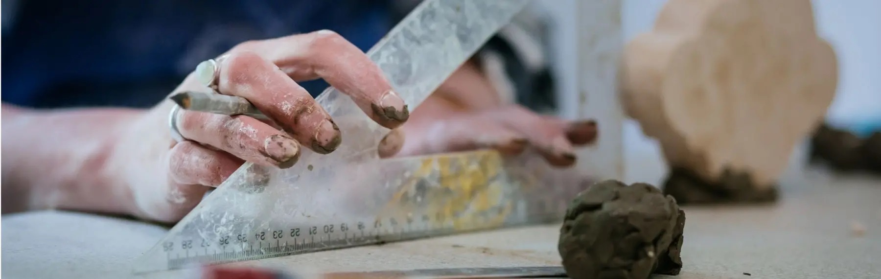 Glendarragh Studios – 25 Years: Sanctuary | Saturday 6 July – Saturday 3 August 2024 | Mermaid Arts Centre | Image: close-up photo of two hands holding a set square, though not to measure, instead to cut into what seems to be a ball of clay; the right hand is holding a pences, and the fingers bear two rings; the fingers have clay on them 