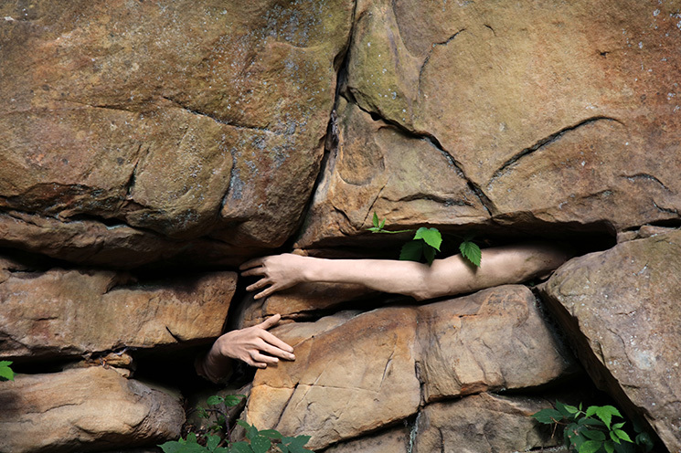 Liane Lang, Fossil Bones, print on Hahnemühle paper | Liane Lang: Deep Time Dip | Saturday 10 August – Sunday 29 September 2024 | Butler Gallery | Image: Liane Lang, Fossil Bones, print on Hahnemühle paper | we see two fairly pale-skinned arms / hands emerging from dark gaps between large boulders – pretty much an impossible situation, it would seem; the arm on the left is mostly just the right hand, while the arm on the right is from shoulder on; the arms cannot be of the same person, because of the separation, but otherwise they looks as though effectively identical; there is also some foliage at gaps between the boulders; most of the image is, in fact, boulders, with little by way of gaps between them 