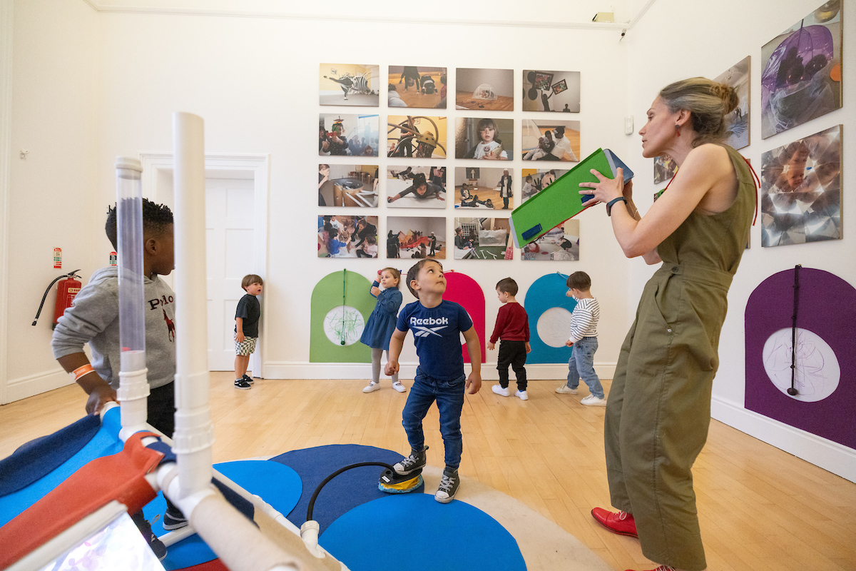 High Expectations: An Interactive Display for Young Children | Tuesday 9 July – Sunday 29 September 2024 | Hugh Lane Gallery | Image: photo of kids plus one adult in a room in (probably) the Hugh Lane; there are photos of kids doing kids stuff in a 4 x 4 grid on the far wall, and close to them we see four kids (aged 3 to 5-ish?) wandering; it looks as though on two of the walls there is sort of automatic-drawing device – sort of a pendulum-like rope with something that makes marks attached to the end, with squiggles on a circular piece of paper just behind the 'pendulum'; two kids nearer us are looking at what the only adult in the room is holding, which is a green construction, shaped somewhat like a large Toblerone bar while still unopened; nearest to us is some sort of plastic and cloth construction of which we only get a partial glimpse 