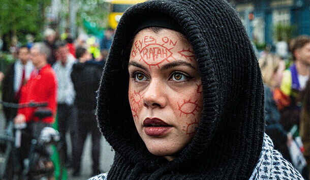 Fragments of Palestine شظايا من فلسطين | from Friday 20 September | RUA RED | Image: photo of someone who is probably at a protest; they’re wearing a thick dark hoodie and loowing past the camera. towards our left; brown eyes, pale-ish skin; the foreheads and cheeks are covered with what are probably eye symbols (though without iris and pupil); the eye on the forehead contains the word RAFAH, with ALL EYES ON written above; the markings are all in red 