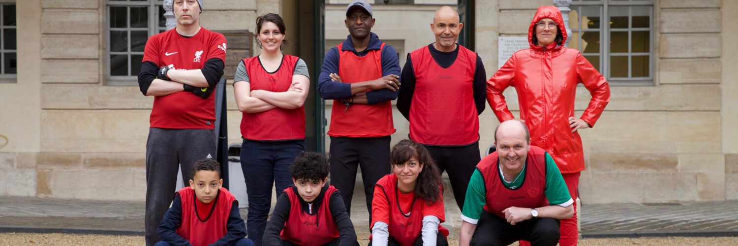 Michael Hanna: Team representing Liverpool | Michael Hanna: Crossing the Park | Saturday 5 October – Saturday 21 December 2024 | CCA | Image: Michael Hanna: Team representing Liverpool | photo of nine people, five standing at the back, four squatting at the front, posing for a team photo; apparent range of gender and age, but all are wearing a very red top – and the figure back right is entirely clad in red, including a hood; some are smiling 