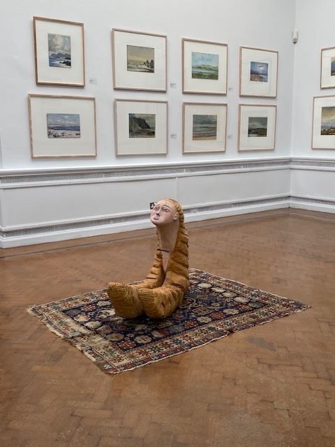 Tina O’Connell | Tina O’Connell: Odysseys of Self | Thursday 12 September – Sunday 6 October 2024 | Limerick City Gallery | Image: Tina O’Connell | photo of a sculpture on an ‘oriental’-style rug in the middle of an exhibition room – we see many landscapes haning on a wall in the background; the sculpture is sort-of mermaid-like – a figure consisting only of head and neck, with the latter disappearing down into two crescent shapes that have extended from the figure’s golden hair; the shapes somewhat resemble large croissants on their sides; the faces is looking up, and looking worried 