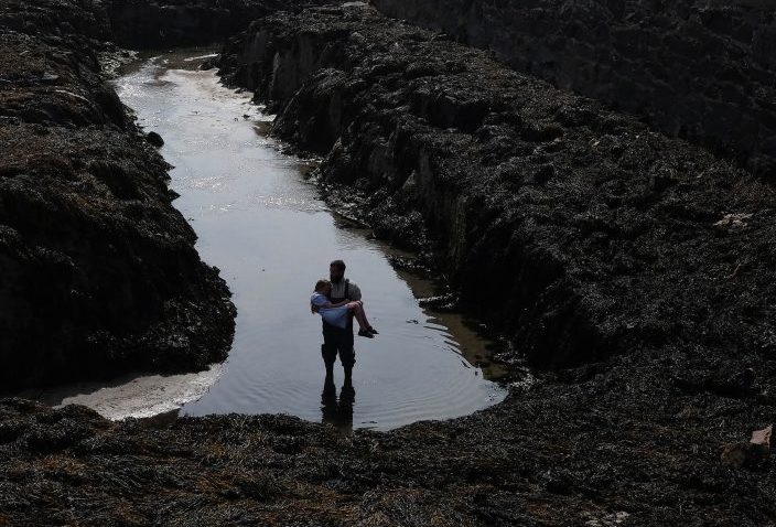 Els Dietvorst (cropped) | Els Dietvorst: ADRIFT | Tuesday 15 October – Friday 29 November 2024 | Wexford Arts Centre | Image: cropped photographic image; form a distance of maybe 20 metres and looking downwards, we see two figures in a water-filled cut in what appears to be a dark bog; the larger figure is holding the smaller above the water 