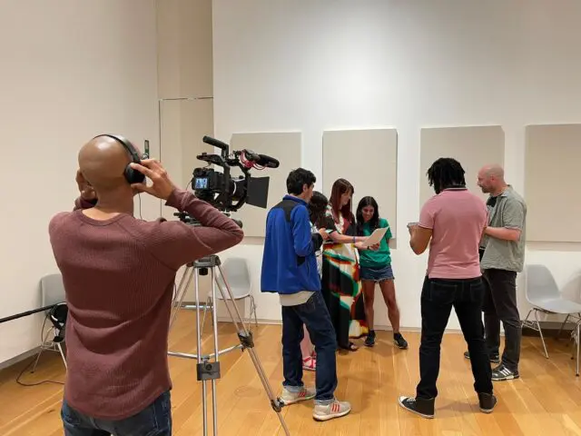 Collaborative Film at Hugh Lane Gallery | Salmagundi: The Project | Saturday 2 November 2024 – Sunday 5 January 2025 | Hugh Lane Gallery | Image: Collaborative Film at Hugh Lane Gallery | photo of a film-making session, presumably in the Hugh Lane; in the foreground is one person, to the left, hands to headphones on a bald head from which light is reflected; in front of the figure – farther from us as the figure’s back is towards us – is a digital film camera with microphone, on a large tripod; farther away, we see six people standing in a circle, facing each other, possibly rehearsing or examining the script; blank canvases appear to be hanging on the wall behind them 