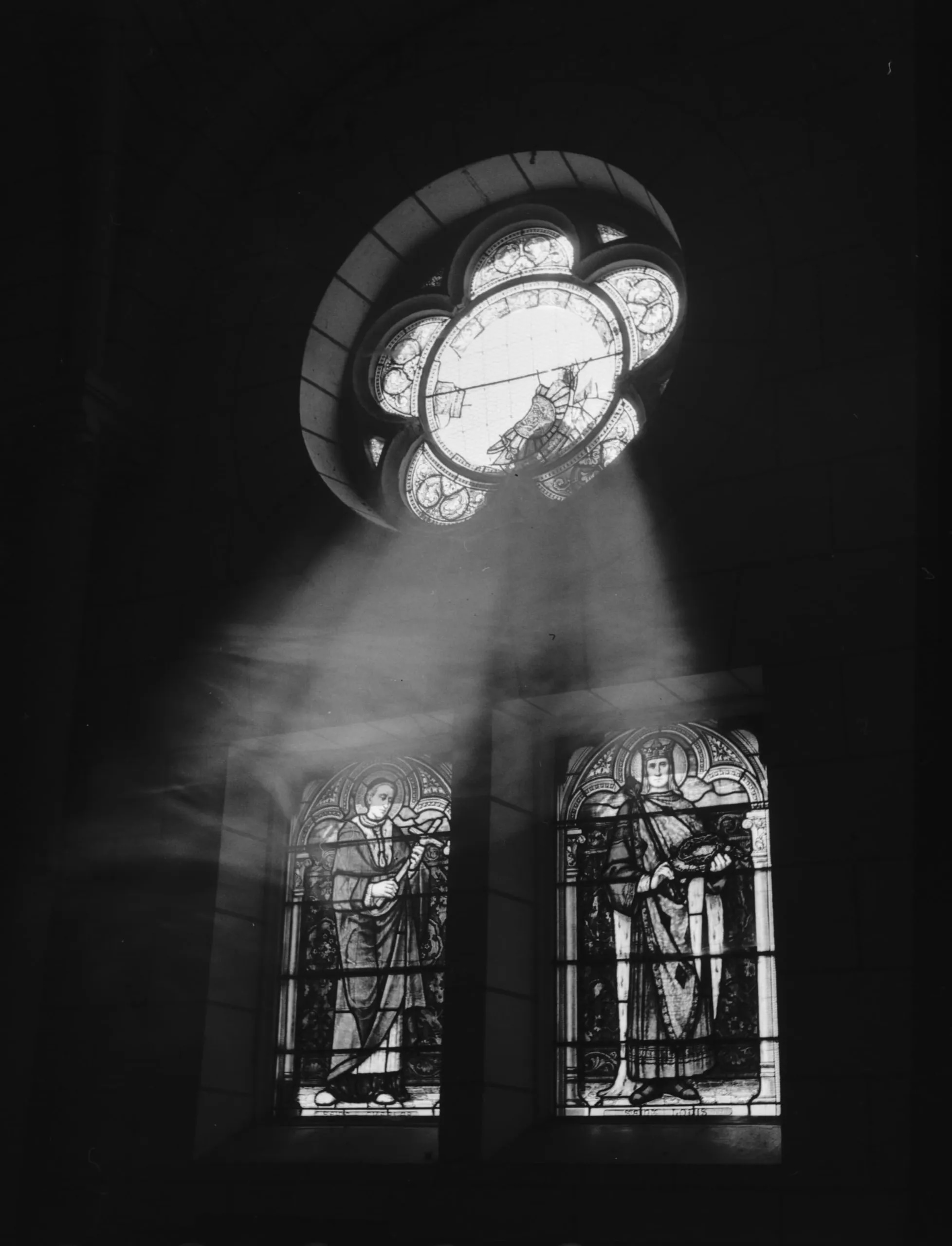 Artem Trofimenko | f.Project: Re-action | Saturday 11 January – Sunday 9 February 2025 | GOMA Gallery of Modern Art | Image: Artem Trofimenko | photo taken in the dark space of a church (presumably; we see three stained-glass windows, viewed slightly from one side; sunlight is screaming in, but it defeated by tye dark after two or three metres; the ‘rose window’ above the other two has five ‘leaves’ around a central circle; it is hard to make out the motifs; the lower two, rectangular windows feature a saint, presumably, in the left one, and a royal character, maybe, in the right one 