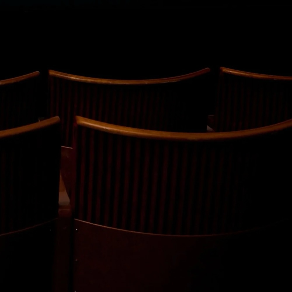 Susan Hiller and Shirin Neshat: Can You Hear Me | Saturday 7 December 2024 – Saturday 1 February 2025 | Golden Thread Gallery | Image: photo / video still, probably (?) of the backs of seats in a cinema; the style is old – wooden seat back, red leather(ette); lighting is from overhead and very restricted, and the seats are looking into a black void 