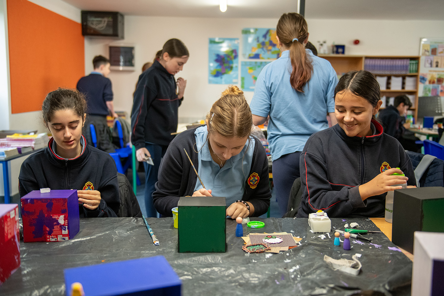 Pupils of CBS, Wexford | Living Arts Project Exhibition | Tuesday 11 March – Tuesday 29 April 2025 | Wexford Arts Centre | Image: Pupils of CBS, Wexford | in the foreground we see three schoolkids, aged around 14?, sitting at a table and working on their art projects, which seem to involve a box shape and paint and small figures; hair tied back , school uniforms in tracksuit version; other kids behind, presumably also engaged in art tasks; maps of Ireland, Europe and the world a wall in the background 