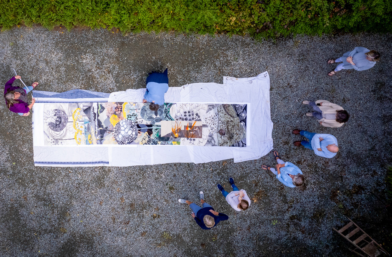 Timelines: Growth and Catastrophe | Friday 30 May – Friday 11 July 2025 | Wexford Arts Centre | Image: photo from above of eight people – presumably the artists; seven are standing, and one is on hands and knees working on a large tapestry spread out on gravelly ground 