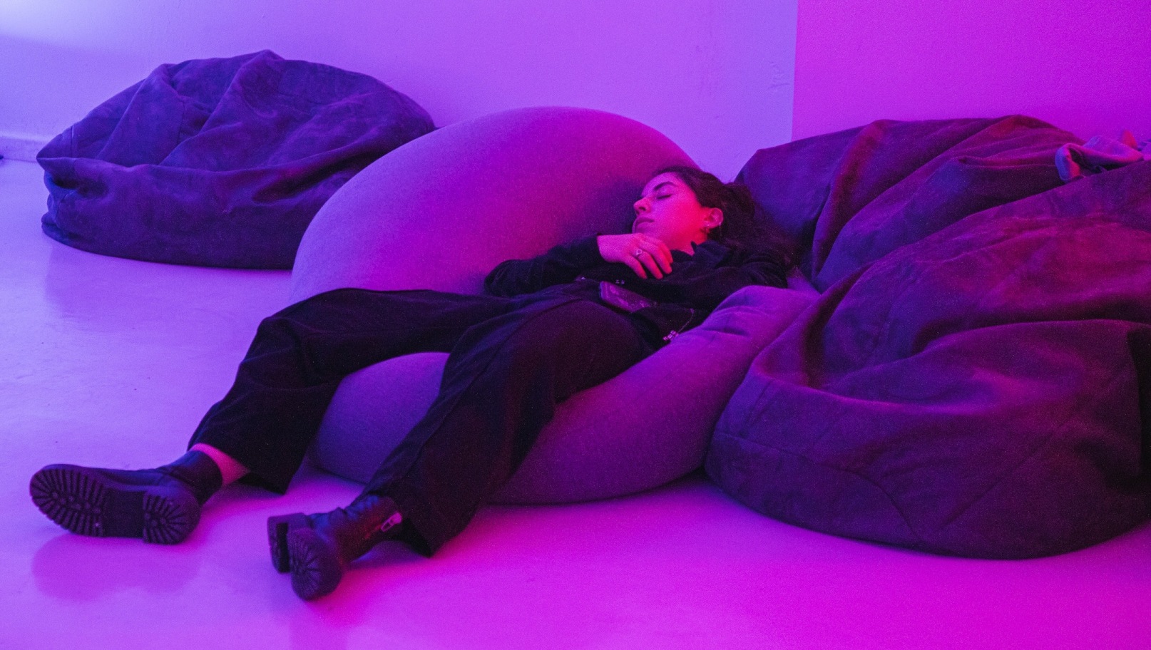 Installation shot; photo by Katie O’Neill | Ciara Barker: Libraries of Rest | Saturday 23 August – Saturday 1 November 2025 | The Dock | Image: installation shot; photo by Katie O’Neill | we see a figure, apparently asleep, lying on a large bean bag in what may be a gallery space; the lighting is mauve; the face and long hair of the figure are more or less centred horizontally in the photo, but about one third from the top; the head is turned a bit to their right, the right hand is just below the chin and we see the right arm, clad in something dark – all the clothese are dark, including the jeans on the legs; black boots stretch towards us and to the left, soles exposed 