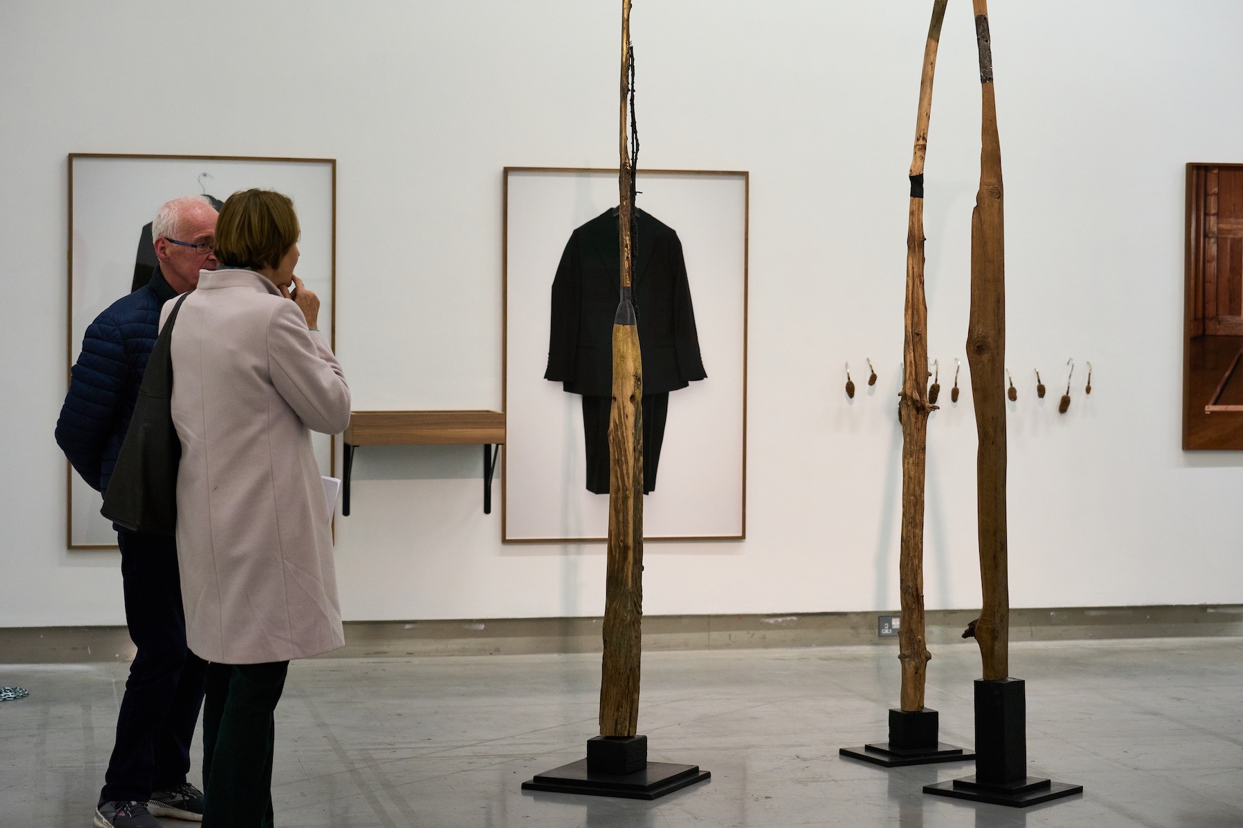 Installation shot, Annual Open Exhibition 2024 | Right Here Right Now – Annual Open Exhibition 2025 | Friday 3 October – Saturday 15 November 2025 | Rua Red | Image: Installation shot, Annual Open Exhibition 2024 | we see two figures, foreground left, contemplating three sculptural pieces that look somewhat like a cross between boat oars and bare trees; on the wall behind we see most of a framed artwork, possibly flat, possibly not, but it looks like a black coat and back trousers, as they might hang together on a hanger, against a white background; the piece is bisected vertically by one of the tree-oar sculpture 