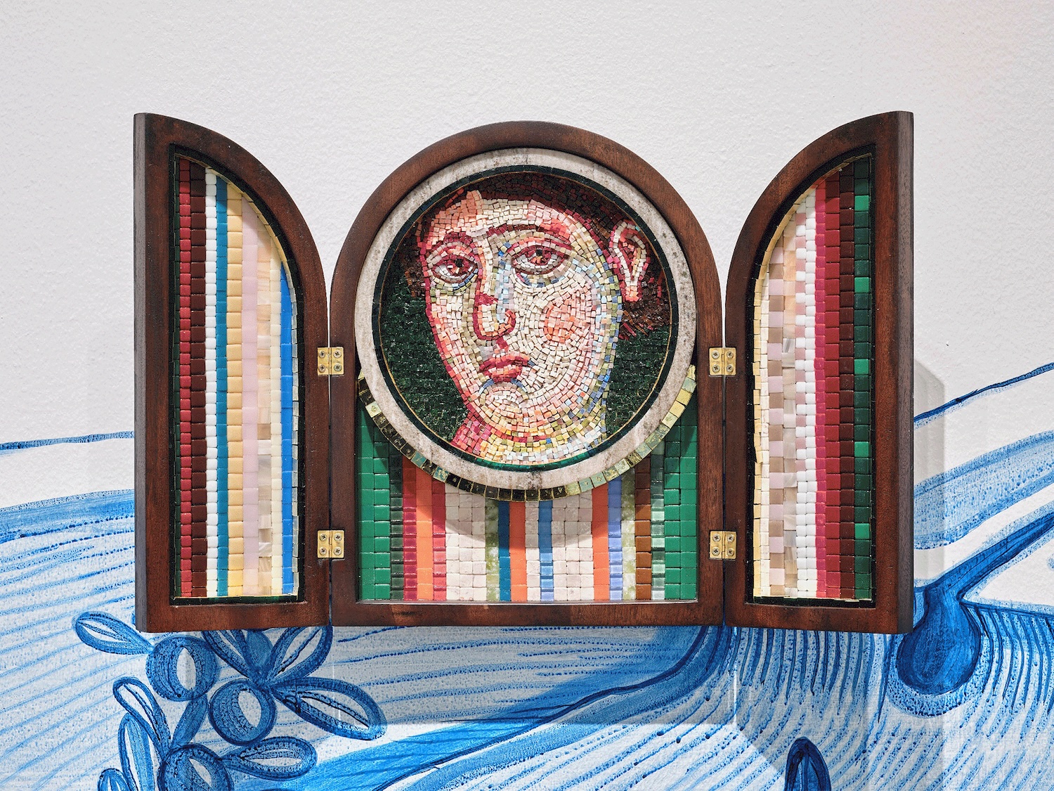 Ursula Burke: Busted Nose, 2024, mosaic glass and custom mahogany frame, 58 x 37cm | Ursula Burke: Siren | Saturday 11 October – Saturday 6 December 2025 | Wexford Arts Centre | Image: Ursula Burke: Busted Nose, 2024, mosaic glass and custom mahogany frame, 58 x 37cm | this is a triptych in a wooden frame which is opened out; shut it would be the shape of a Roman arch, open it is two half-arches and a central arch; the semicircle of that arch becomes a circle which holds a mosaic of a face looking out at us quizzically; the lower part of the central panel and other panels in their totality are covered with colourful mosaic – patterned except for the face; the face has a red scar half way up the nose 