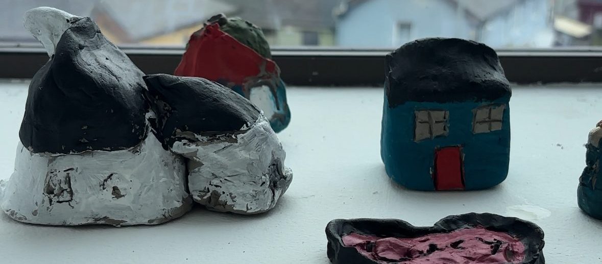 Harvest | Monday 29 September – Saturday 11 October 2025 | Uillinn: West Cork Arts Centre | Image: a photo of some painted clay objects made by the kids; shapes on the left may just be penguins; a shape beyond the penguins may be a house, and the shape on the right is definitely a house, with blue-painted walls, a black roof and a red door; there is a partial view of what may be a painted clay plate of some sort; through a window behind all this, we get a glimpse of the town 