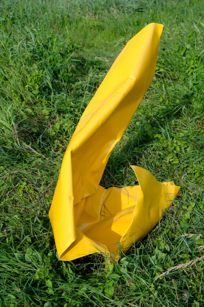 Padraig Cunningham, Caoimhe Kilfeather, Nollaig Molloy: Extract | Friday 27 January  – Saturday 1 April 2023 | Roscommon Arts Centre | Image: photo of what seems to be a large piece of yellow plastic, in a green, grassy field; the plastic is fairly rigid, and has been bent into a sort of leaning L, with its foot capturing a small pool of water. 