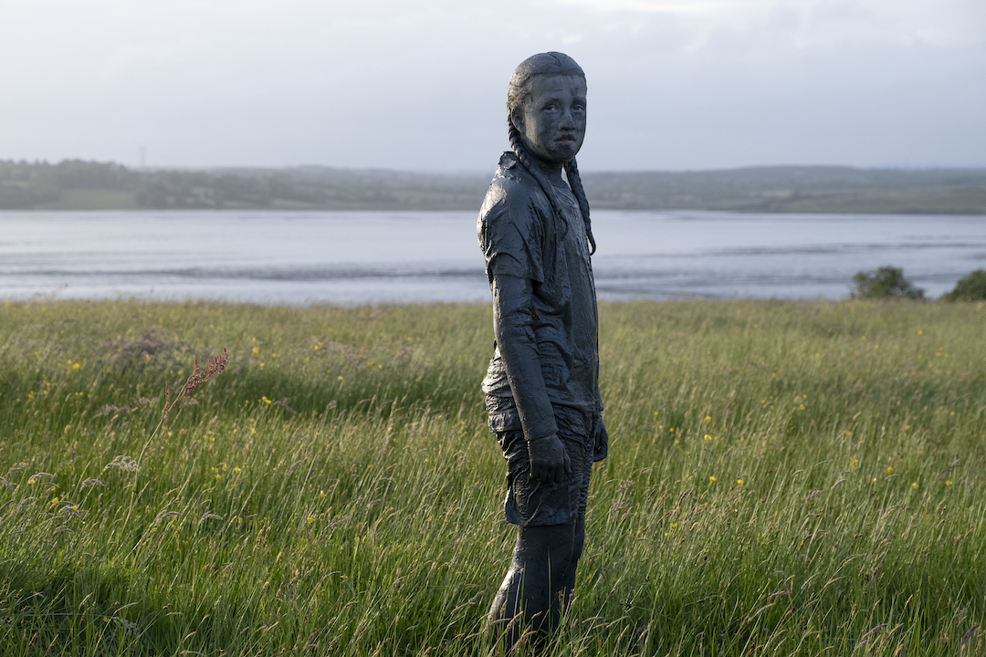 Eve, 2022, photograph by William Bock, courtesy the artist | River Residencies | Friday 17 February  – Saturday 8 April 2023 | Ormston House | Image: Eve, 2022, photograph by William Bock – the figure is apparently covered from head to toe in what looks like mud or crude oil; behind is a riverine landscape 