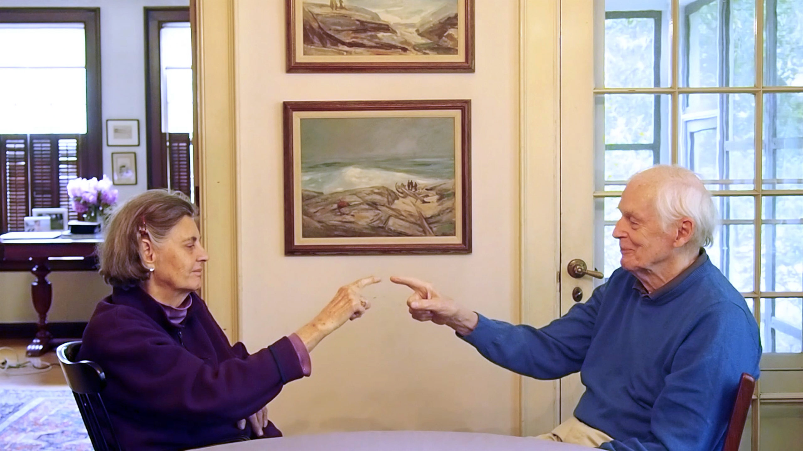 Sarah Pierce: Scene of the Myth | Friday 24 March  – Sunday 3 September 2023 | IMMA | Image: Photograph. We see a somewhat elderly couple sitting at a table in a domestic location; each has their right arm raised and their index finger pointing right at the other’s index finger, which are almost touching; there are paintings on the wall – landscapes – right behind where the fingers almost meet; to the left, behind, we see flowers on a table and partial glimpses of two large, partially shuttered windows; at the right of the photo there is a door leading into what may be a conservatory, with glimpses of a possible garden outside 