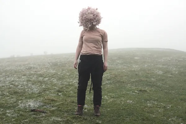 image from the artist's website | Joanna Hopkins: Fruity Bodies | Saturday 25 February  – Sunday 9 April 2023 | GOMA Gallery of Modern Art | Image: photo of female standing in a misty field; wearing black jeans and a pink T shirt, with a fluffball where the head should be 