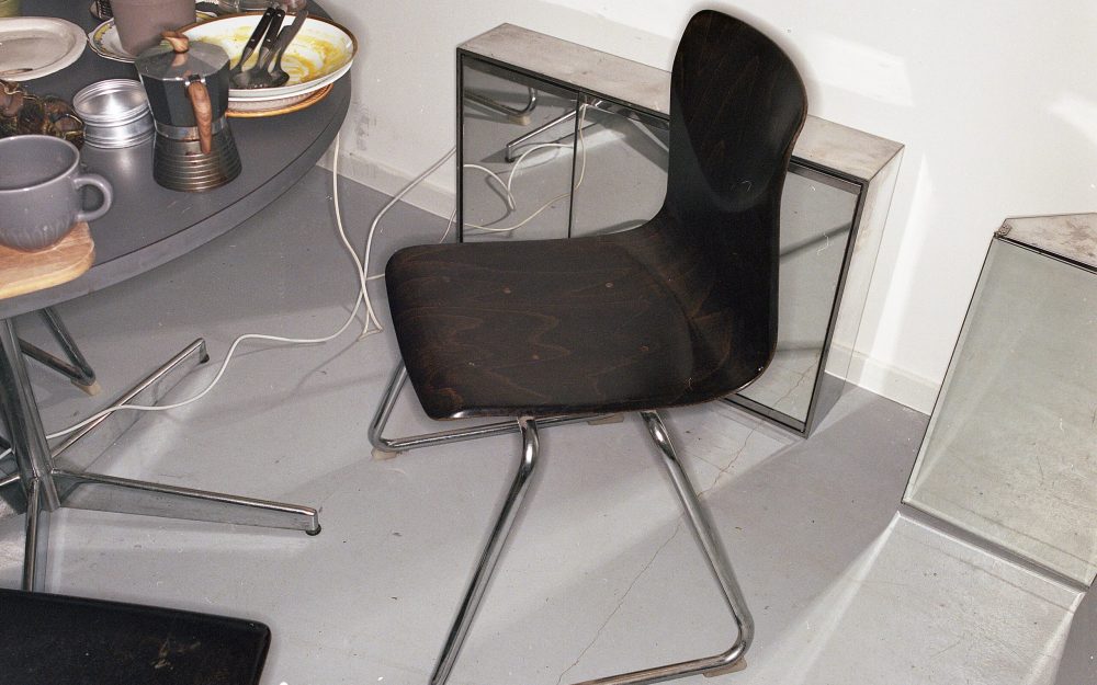 Áine Mac Giolla Bhríde: objects as buoys in the life acted | Saturday 15 April  – Saturday 13 May 2023 | Regional Cultural Centre | Image: mute-coloured photo (?) of a table, chair, mirrored cabinet on the floor, in what looks like an office or workspace; breakfast (?) remains on table 