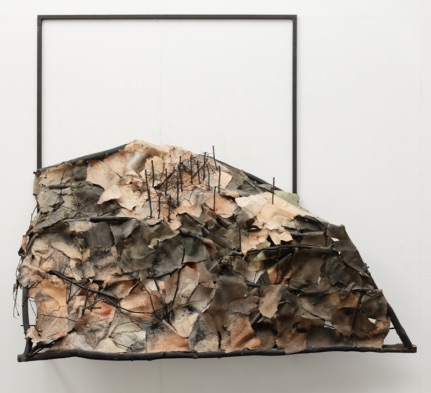 Art and the Anthropocene: Being in and Apart from Nature | Friday 3 March  – Saturday 6 May 2023 | Garter Lane Arts Centre | Image: on a metal or maybe twig frame sticking out from the wall, we see what looks like a hillside made of discoloured cloth or paper; bare black tree trunks are mostly clustered towards the top of the hill; there is a square metal frame behind the hill 