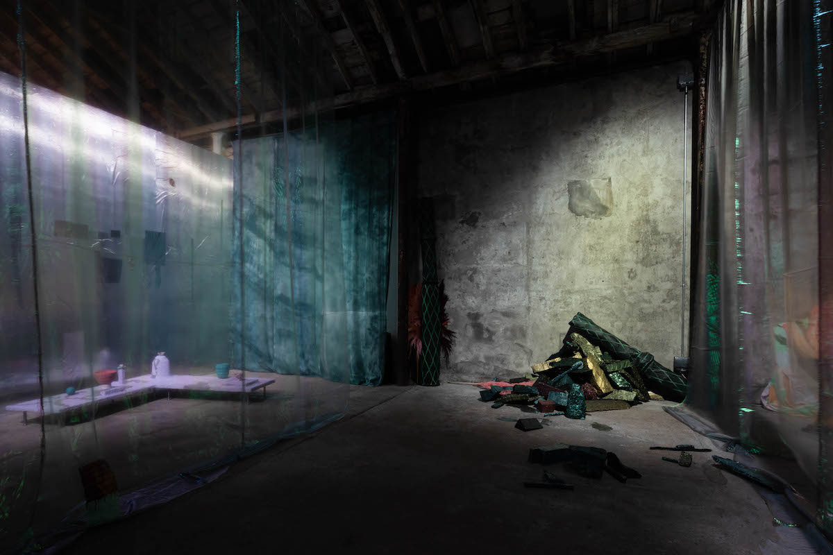 Eleanor McCaughey and Lucy Sheridan, Pay No Attention to that Man Behind the Curtain, Installation view Faux rubble, polystyrene, solar dust and faux columns, paper mache, acrylic paint and collage, 2023 | PORTALS | Saturday 29 April  – Thursday 22 June 2023 | Luan Gallery | Image: Eleanor McCaughey and Lucy Sheridan, Pay No Attention to that Man Behind the Curtain, Installation view Faux rubble, polystyrene, solar dust and faux columns, paper mache, acrylic paint and collage, 2023 – looks like the interior of large shed with concrete floor and walls; large transparent plastic separators between rooms; rolls of patterned material stacked against the wall in one space; in a second, ceramic objects are set out on a very low L-shaped table 