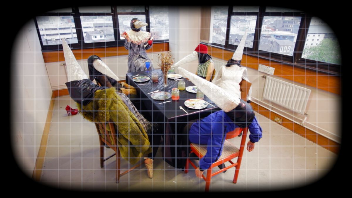 Natasha Bourke, Concrete Keys (still), 2022. Video, colour, sound, 120 minutes. Courtesy of the artist | Natasha Bourke: Concrete Keys | Saturday 22 April  – Saturday 20 May 2023 | SIRIUS | Image: Natasha Bourke, Concrete Keys (still), 2022. Video, colour, sound, 120 minutes. Courtesy of the artist – video still of six people at a table set for eating, though in an otherwise bare office room; each participant is wearing what looks like a black plastic bag on their heads, and a metre-long white 'beak' which looks like a dunce's hat 