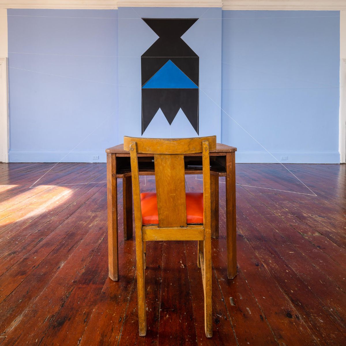 View of Patrick Ireland, aka Brian O'Doherty, HCE Redux, 2004 (remade 2023), SIRIUS, 2023. Paint, cord, table, chair, typed paper. Dimensions variable. Courtesy of The Estate of Brian O’Doherty. Photograph: John Beasley | SIRIUS Summer School: Brian O’Doherty: Reading Time | Tuesday 6 June  – Saturday 10 June 2023 | SIRIUS | Image: View of Patrick Ireland, aka Brian O'Doherty, HCE Redux, 2004 (remade 2023), SIRIUS, 2023. Paint, cord, table, chair, typed paper. Dimensions variable. Courtesy of The Estate of Brian O’Doherty. Photograph: John Beasley – in a large room: in the foreground a the back of a chair at a small table, both wooden; on the wall some 5 metres (?) further into the room, a geometrical pattern on the pale-blue wall; some strings link the pattern to the floor 