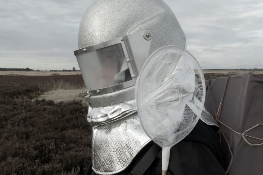 Hanne Nielsen and Birgit Johnsen: Particles | Friday 28 April  – Saturday 24 June 2023 | Ormston House | Video still: against a boggy landscape, someone in a semi-beekeeper, semi-astronaut helmet is apparently holding a butterfly net (we only see head and shoulders 