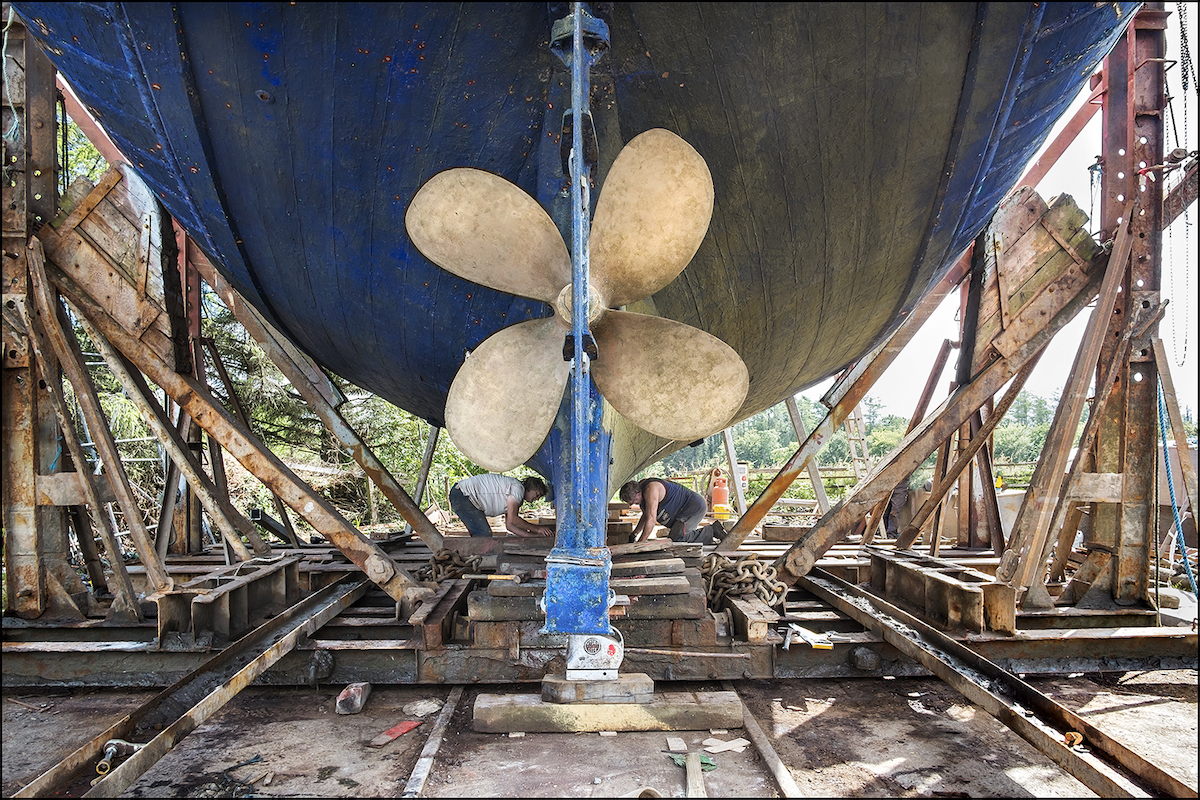 Kevin O’Farrell: Hegarty’s Boatyard | Saturday 13 May  – Saturday 10 June 2023 | Uillinn: West Cork Arts Centre | Image: Kevin O'Farrell: Repairing the keel on a 56’ wooden trawler, Hegarty’s Boatyard, 2014 – under what seems a huge hull we see two men working on a boat's keel; the large propellor and (possibly) rudder are closest to us; the hull is supported by a complex arrangement of girders short, thick beams  