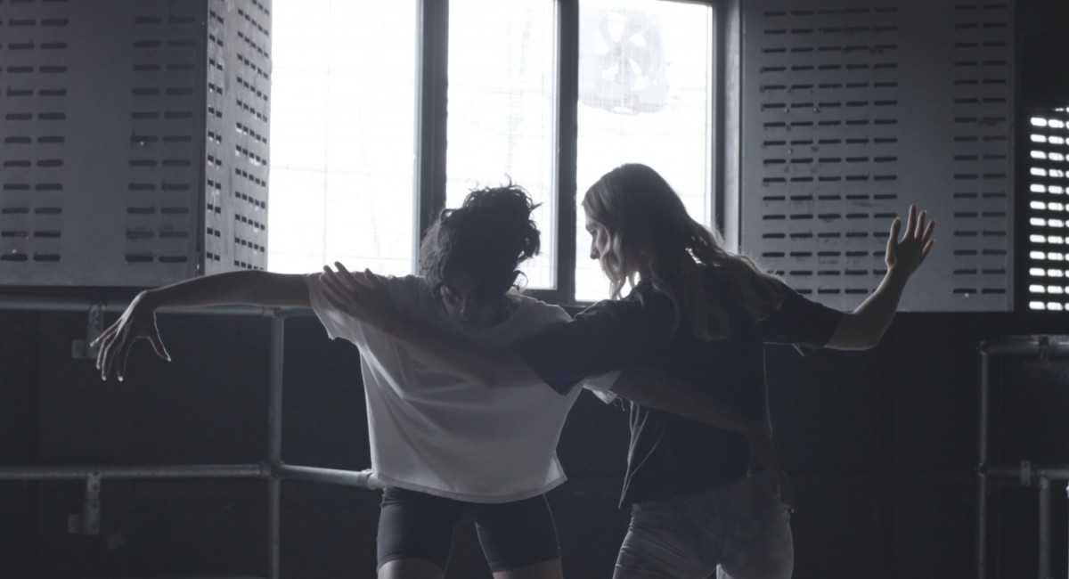 Amanda Rice, The Flesh of Language (still), 2023. Video, colour, sound, 12 minutes. Courtesy of the artist | Amanda Rice: The Flesh of Language | Saturday 27 May  – Saturday 24 June 2023 | SIRIUS | Image: Amanda Rice, The Flesh of Language (still), 2023. Video, colour, sound, 12 minutes. Courtesy of the artist – the video still shows two figures facing each other, arms outstretched and overlapping; possible a dance move; window behind with slitted shutters 