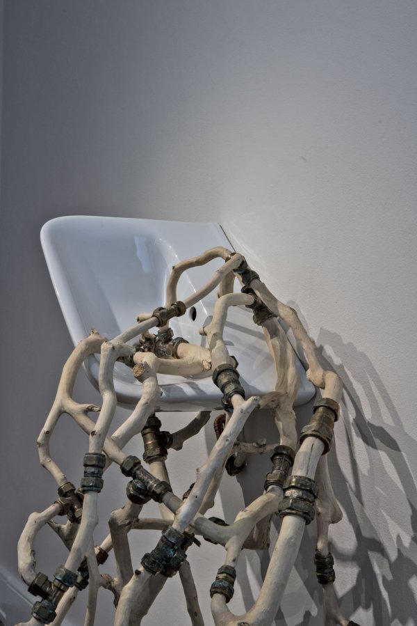 Photo credit: Paul McCarthy | Katherine Sankey: an atom-bomb in each morsel of life | Thursday 15 June  – Tuesday 5 September 2023 | The LAB | Image: Photo credit: Paul McCarthy – we see a white ceramic sink attached to a wall, viewed from above or tilted; coming from the near side of the sink there is what looks like bones, but they are joined together in an organic mesh by metal connectors 