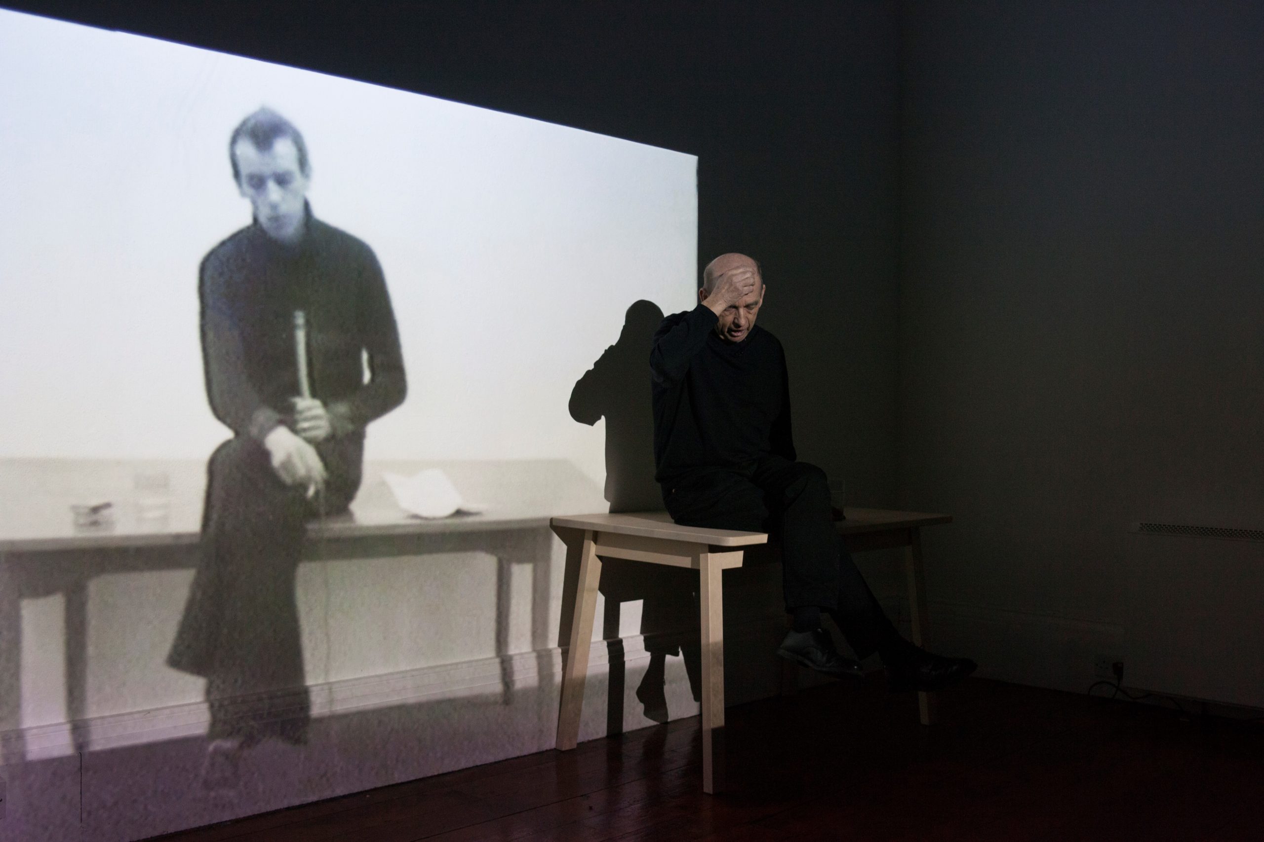 © Kevin Atherton | Image courtesy of the artist and Miranda Driscoll and Sirius Arts Centre, Cobh | Kevin Atherton: In Two Minds | Friday 26 May  – Sunday 8 October 2023 | IMMA | Image: © Kevin Atherton | Image courtesy of the artist and Miranda Driscoll and Sirius Arts Centre, Cobh – we see the artist, dressed in black and with right hand raised to forehead, sitting on a table and caught in the light from a projector showing what is probably an image the artist when younger, apparently holding a microphone in one hand and smoking with the other 