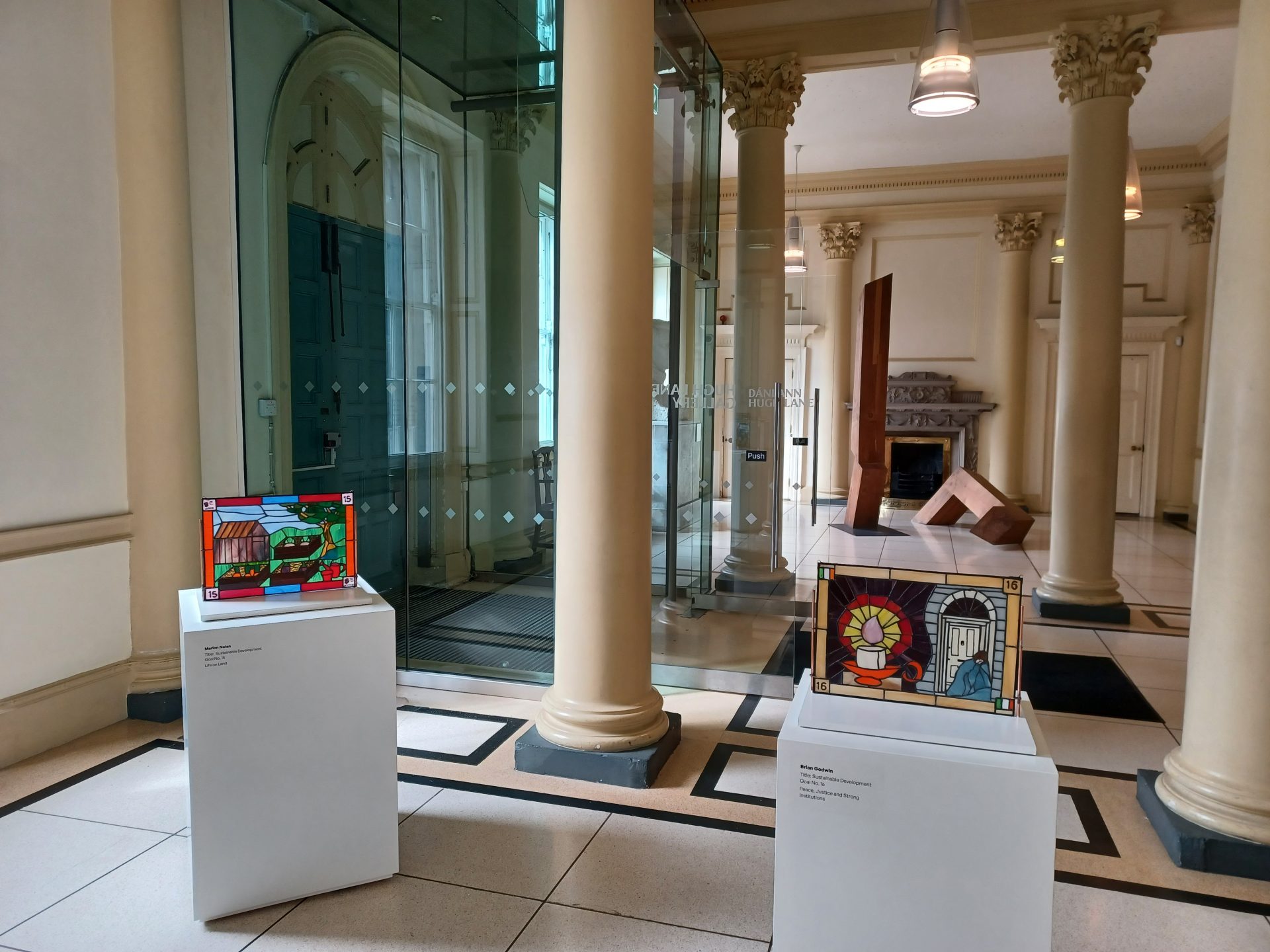 Solder: Global Connection through Art | Thursday 18 May  – Sunday 11 June 2023 | Hugh Lane Gallery | Image: photo of two stained-glass works, each about A2 size (?) on plinths in the entrance hall of the Hugh Lane; the closer work depicts a  lit candle in candle-holder on the left, and a rough-sleeper in a Georgian doorway on the right; the farther work suggests a rural landscape, or – more likely – an allotment, with plants growing in raised beds 