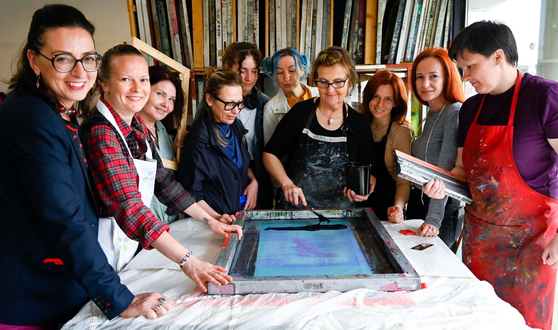 InFlight_OnLanding | Tuesday 20 June  – Tuesday 27 June 2023 |  | Image: photo of artist Varvara Keidan Shavrova and ten refugees gathered around as ink is applied to a silkscreen mesh for printing onto what appear to be white fabric 