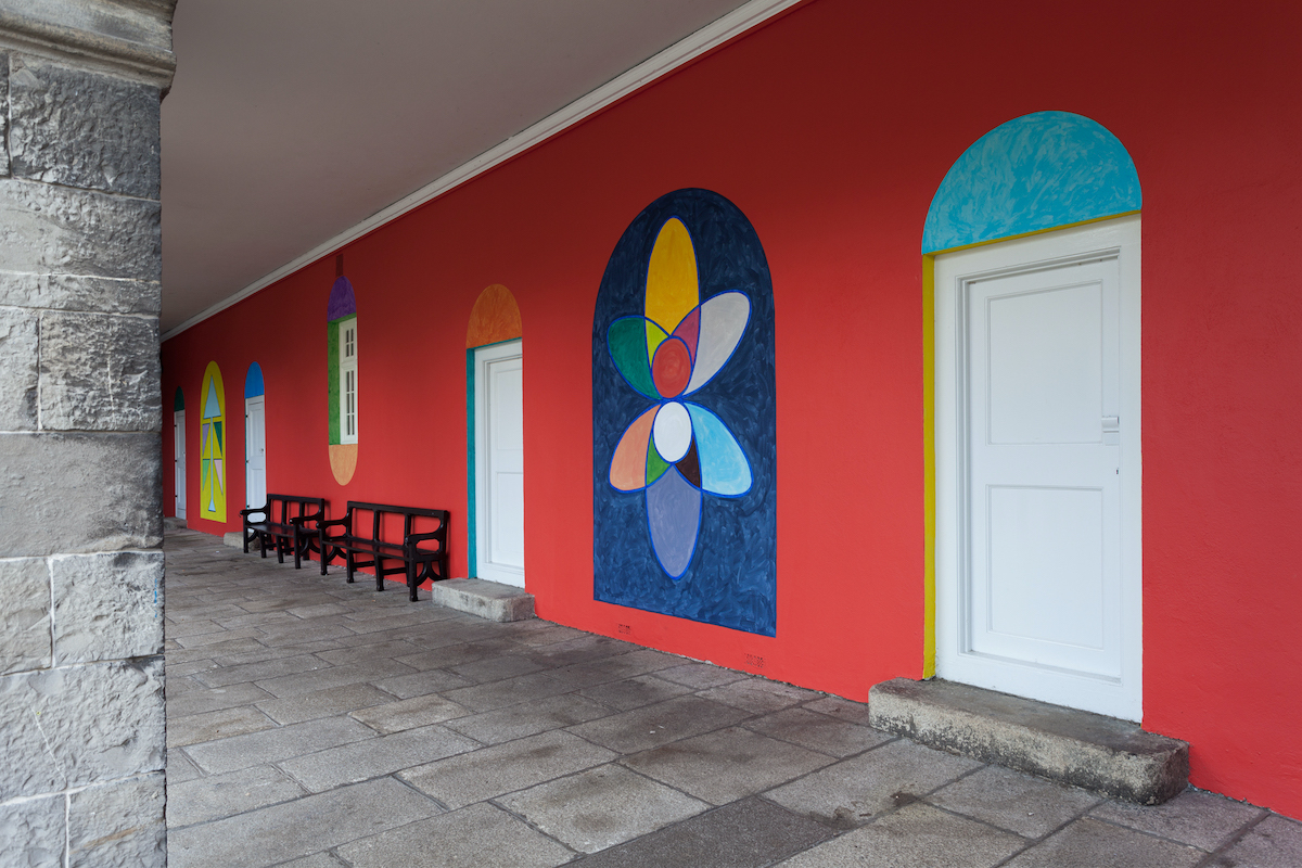 Navine G. Dossos: Kind Words Can Never Die | Thursday 28 July 2022  – Tuesday 30 July 2024 | IMMA | Image: photo of one of the colonnades in the IMMA courtyard, taken at an angle so that we see a number of interventions by the artist; the wall itself has been painted red, except where the artist has intervened with other colours in various shapes; each (white) door along the wall has above it a brightly coloured semicircle, very much like the fanlights above doors in Georgian parts of Dublin, though without any of the fan detail; between the doors, including around the only window, are large shape painted shapes in mostly bright colours; for example, the large shape nearest the camera is a dark-blue rectangle with a rounded top, onto / in which is a multicoloured shape which looks roughly like it is showing the paths of electrons around an atom 
