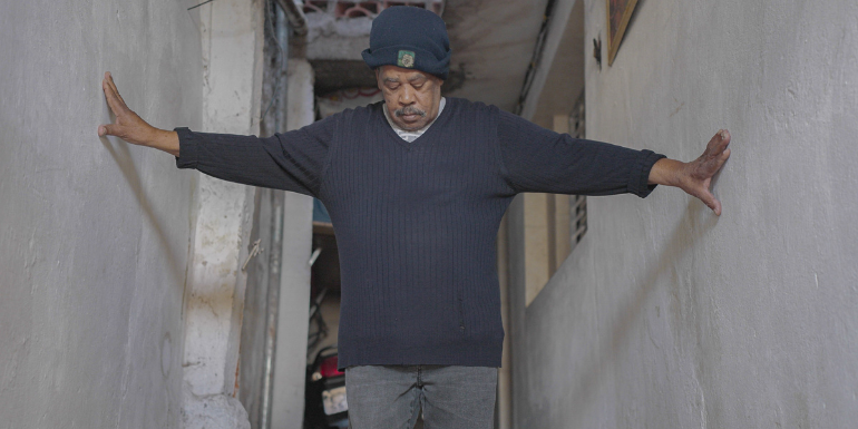 Robert Chase Heishman: The House that Valdemar Built, film still | Brian Maguire, James Concagh, Robert Chase Heishman: Then I Laid the Floor | Saturday 15 July  – Tuesday 31 October 2023 | Triskel Arts Centre | Image: Robert Chase Heishman: The House that Valdemar Built, film still – we see a figure in a grey-blue woolly hat and grey-blue sweater looking downwards and with arms outstretched touching what may be just-plastered walls in what may be a house under construction 