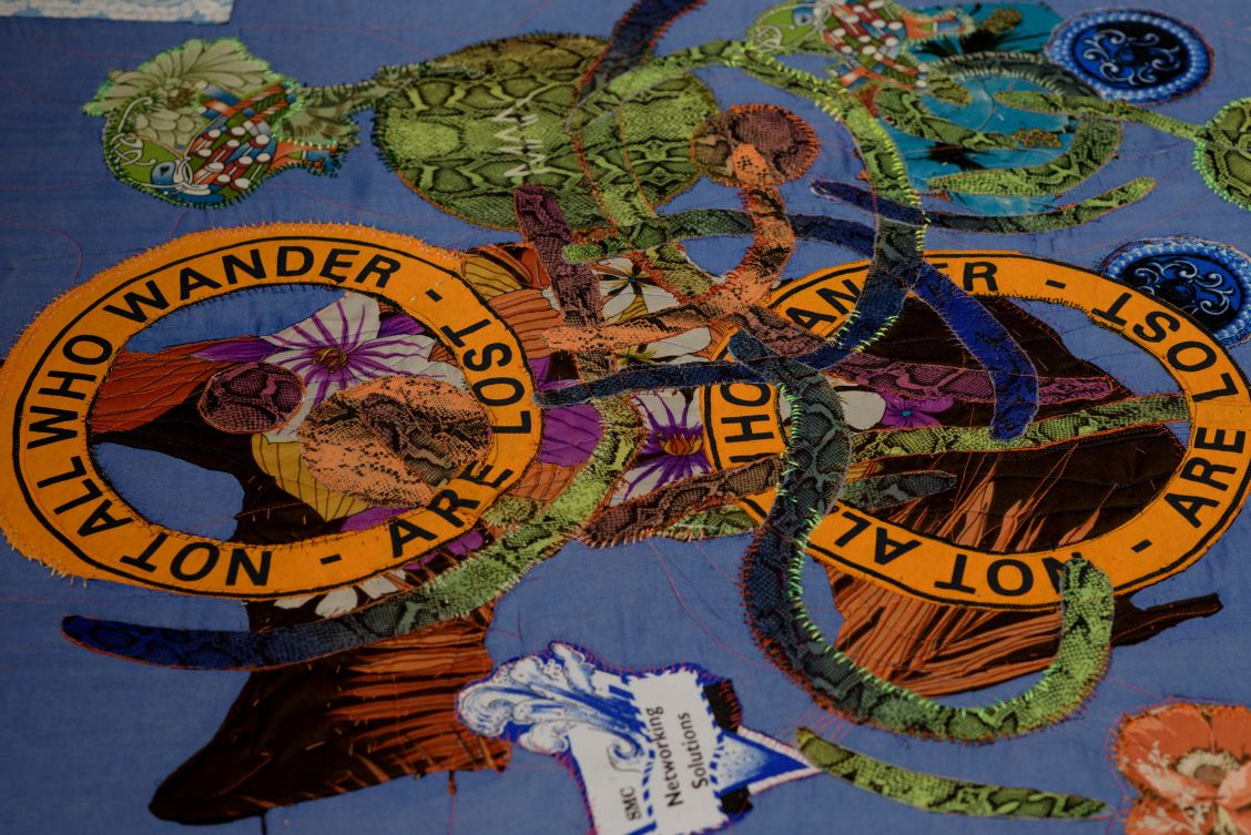 Those Who Wander, Sibyl Montague, 2021, hand stitched & embroidered found textiles. Courtesy of the artist. Photo by Louis Haugh. | Marie Farrington, Sibyl Montague, Laura Ní Fhlaibhín and Liliane Puthod: Hammerheads | Saturday 1 July  – Saturday 16 September 2023 | Solstice Arts Centre | Image: 'Those Who Wander', Sibyl Montague, 2021, hand stitched & embroidered found textiles. Courtesy of the artist. Photo by Louis Haugh.  – various pieces of coloured fabric stitched onto a pale, grey-blue background; two of the stitched-on pies are circles of text on an orange background; they read “Not all who wander are lost” 